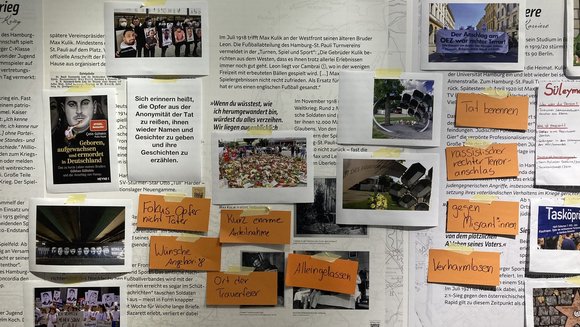 Bilder, Zitate und beschriftete Kärtchen kleben an einer Wand. Auf den Bildern sind trauernde Menschen und Denkmäler.