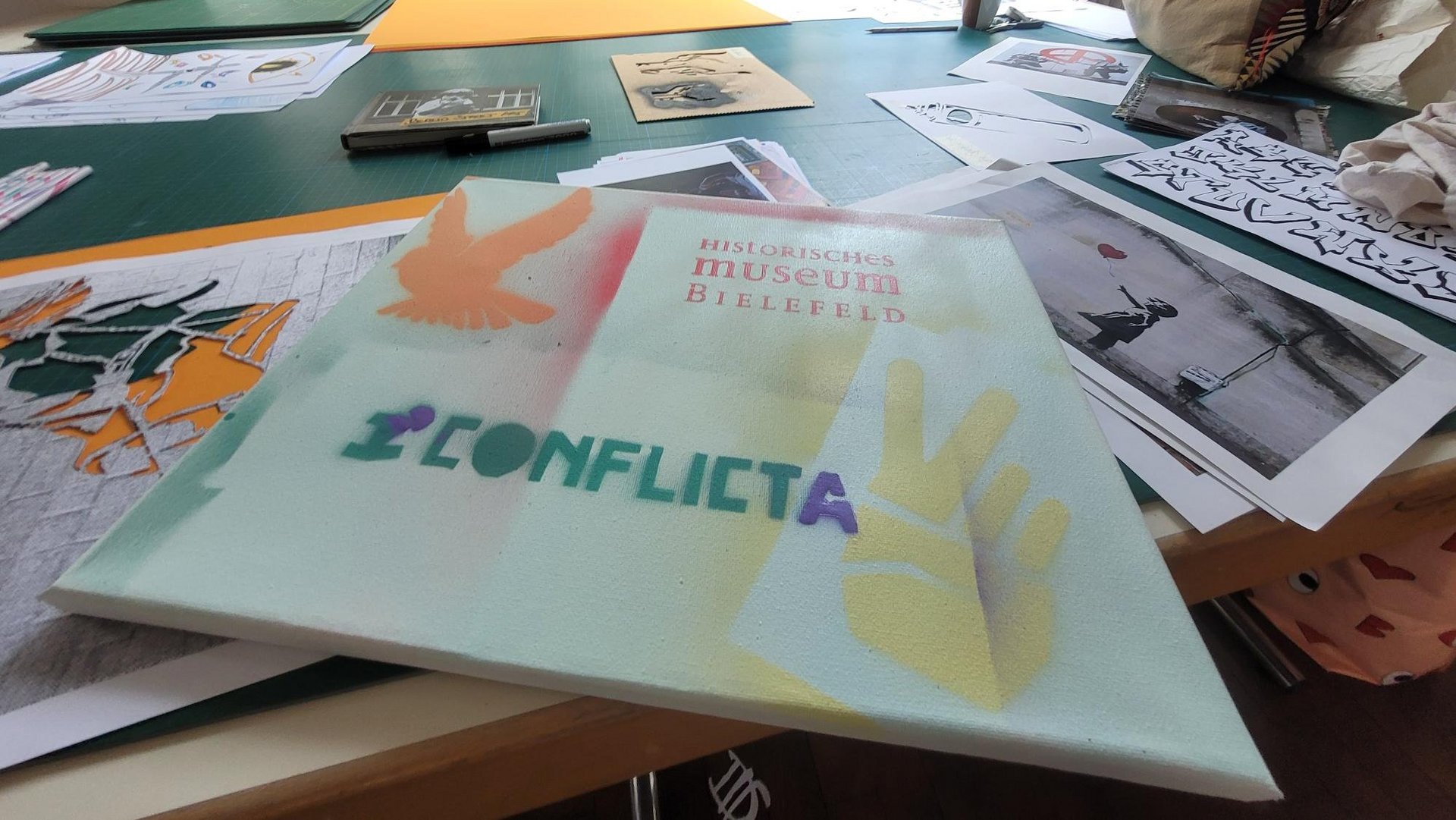 Ein Tisch mit Bildern darauf. Auf eine Leinwand ist „Historisches Museum Bielefeld ConflictA“ gesprayed.