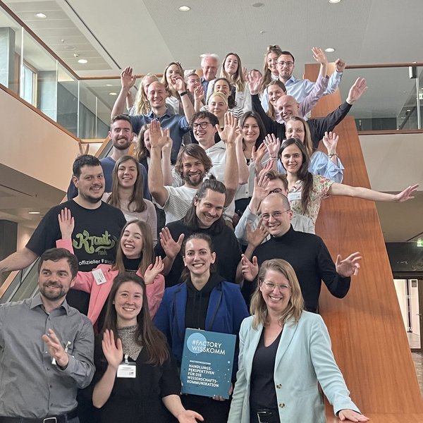 Projektbild Hochschulwettbewerb: Foto mit etwa 25 Menschen, die von einer Treppe winken. Eine Frau in der ersten Reihe hält eine Mappe mit der Aufschrift "#FACTORY WISSKOMM".