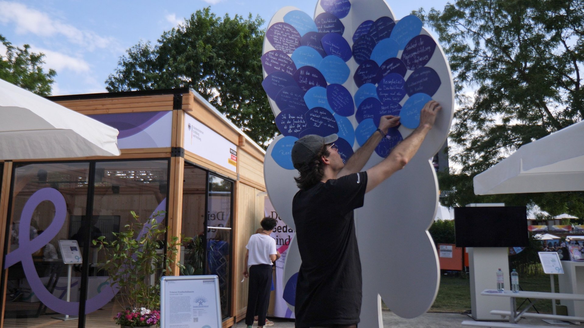 Eine junge Person heftet lila und blaue Sprechblasen an einen großen violetten Baum aus Pappe