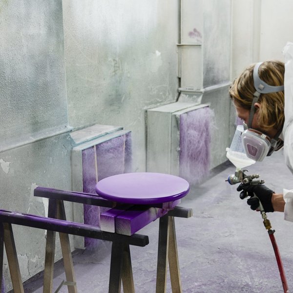 Eine Person sprüht lila Farbe auf eine runde Platte. Sie trägt einen Maleranzug und eine Schutzmaske. 
