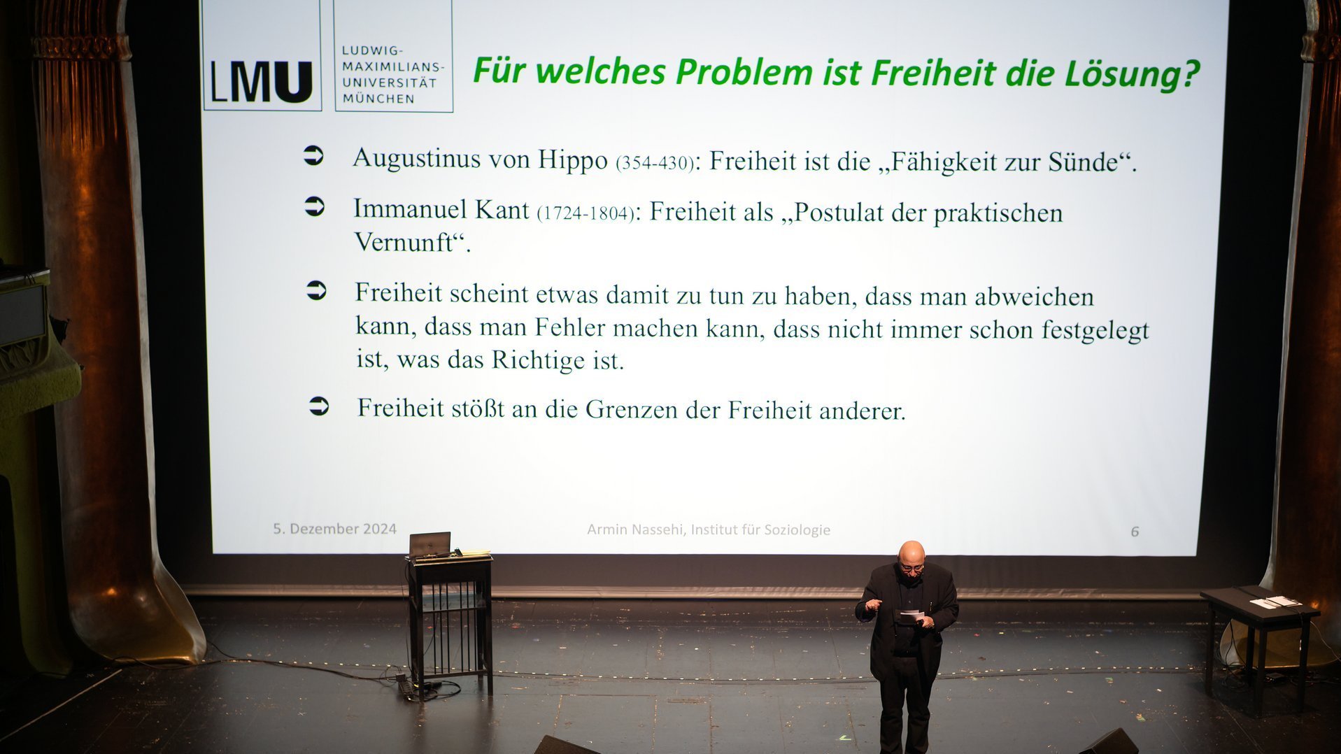 Soziologieprofessor Armin Nassehi hält einen Vortrag auf der Bühne vor Publikum, im Hintergrund eine Folie mit philosophischen Gedanken zu Freiheit.