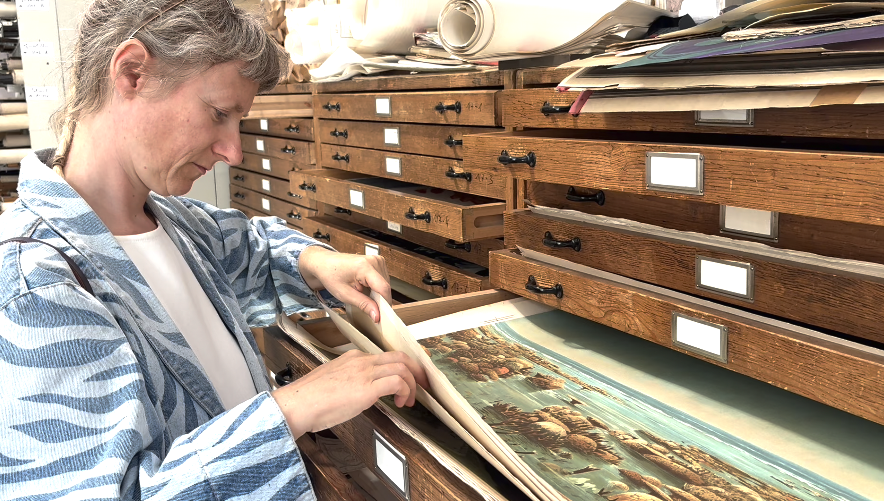 Line Krom steht an einem Grafikschrank, eine Schublade ist geöffnet, sie hebt ein darin liegendes Gemälde an.