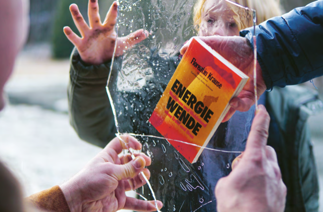Eine Hand hält das Buch "Energiewende" gegen ein Stück einer Glasscheibe, die eine andere Person mit zwei Händen hält.