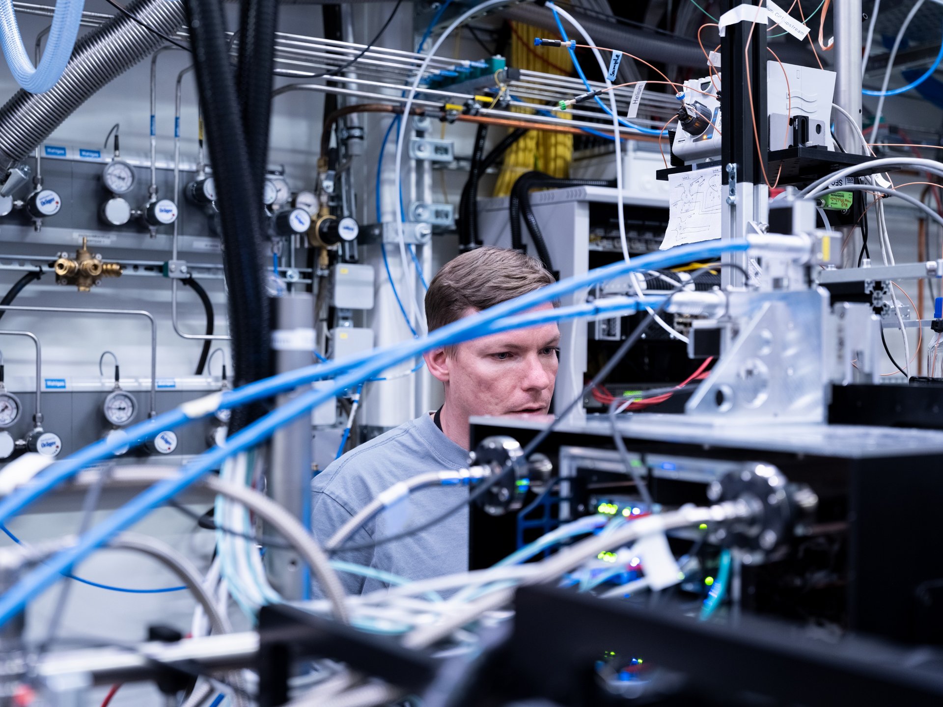 WJ25_idw_European XFEL.jpg Ein junger Mann sitzt zwischen vielen technischen Geräten. Er schaut konzentriert auf die Geräte. Hinter ihm sind weitere Rohre und Messanzeigen zu sehen.
