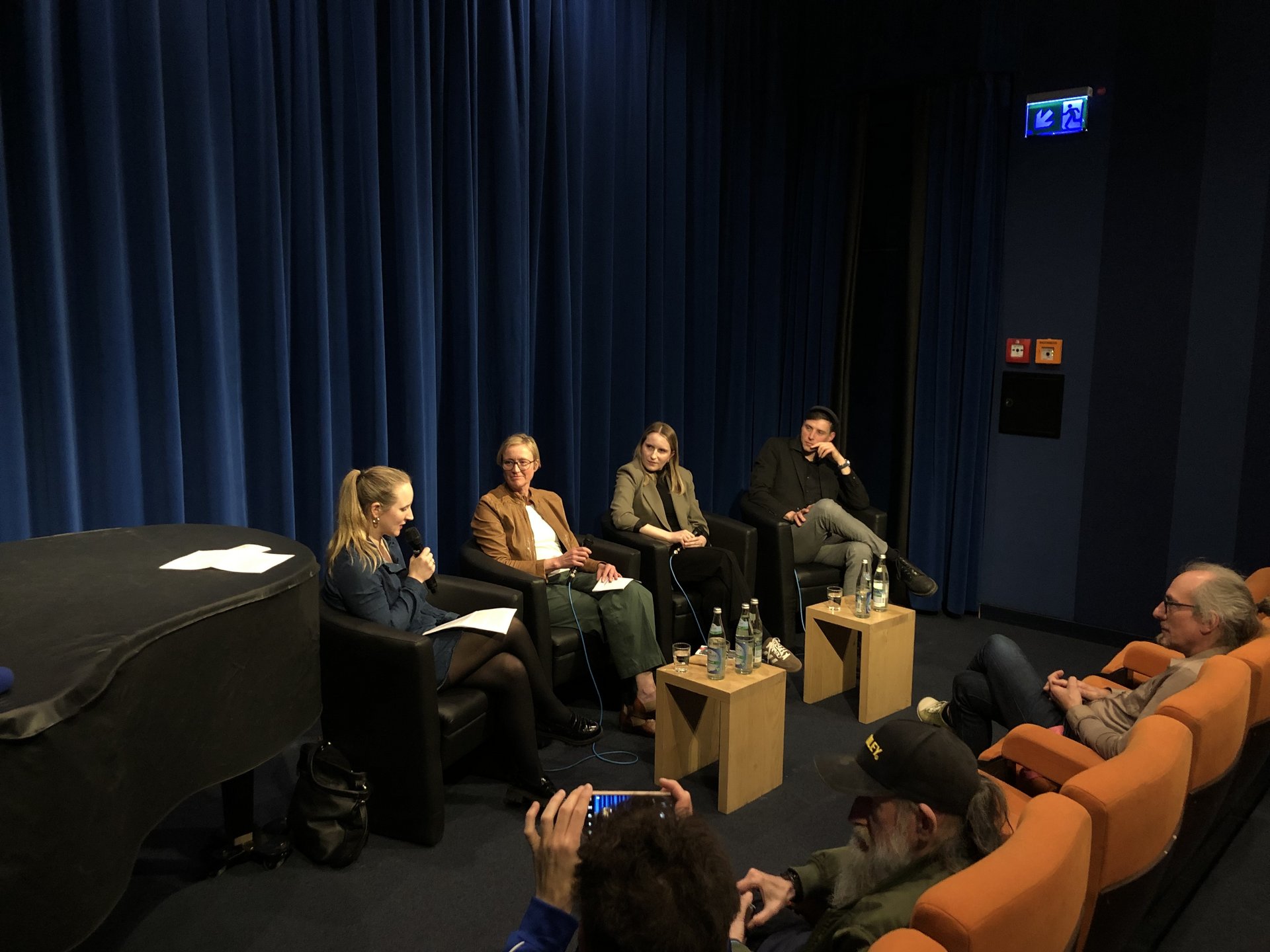 Vier Personen sitzen in einem Kinosaal vor Publikum, eine Moderatorin spricht in ein Mikrofon.