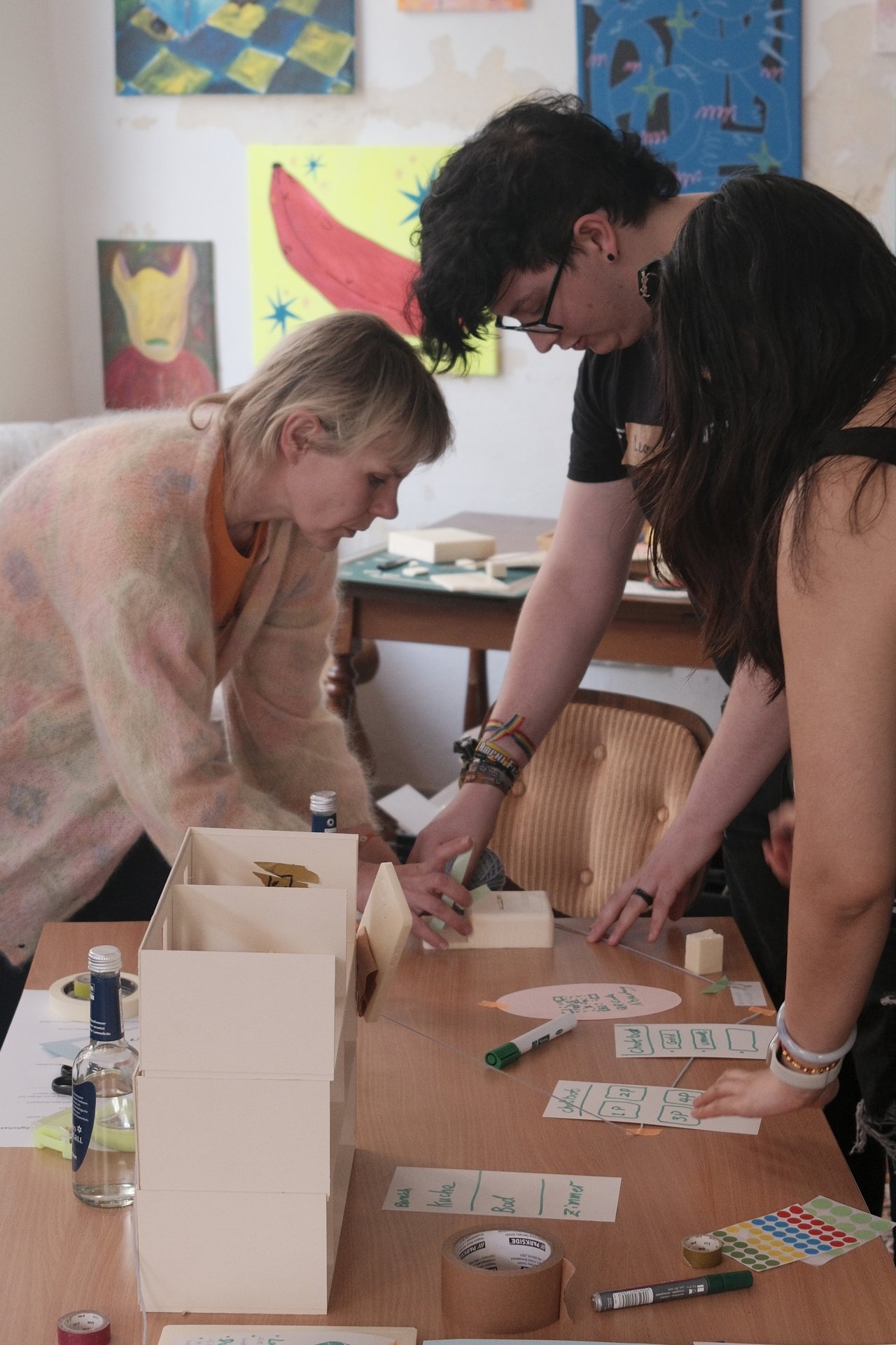 Drei Personen gemischten Alters stehen über einen Tisch gebeugt. Auf dem Tisch befinden sich ein Modellhaus und verschiedene Schreibutensilien.