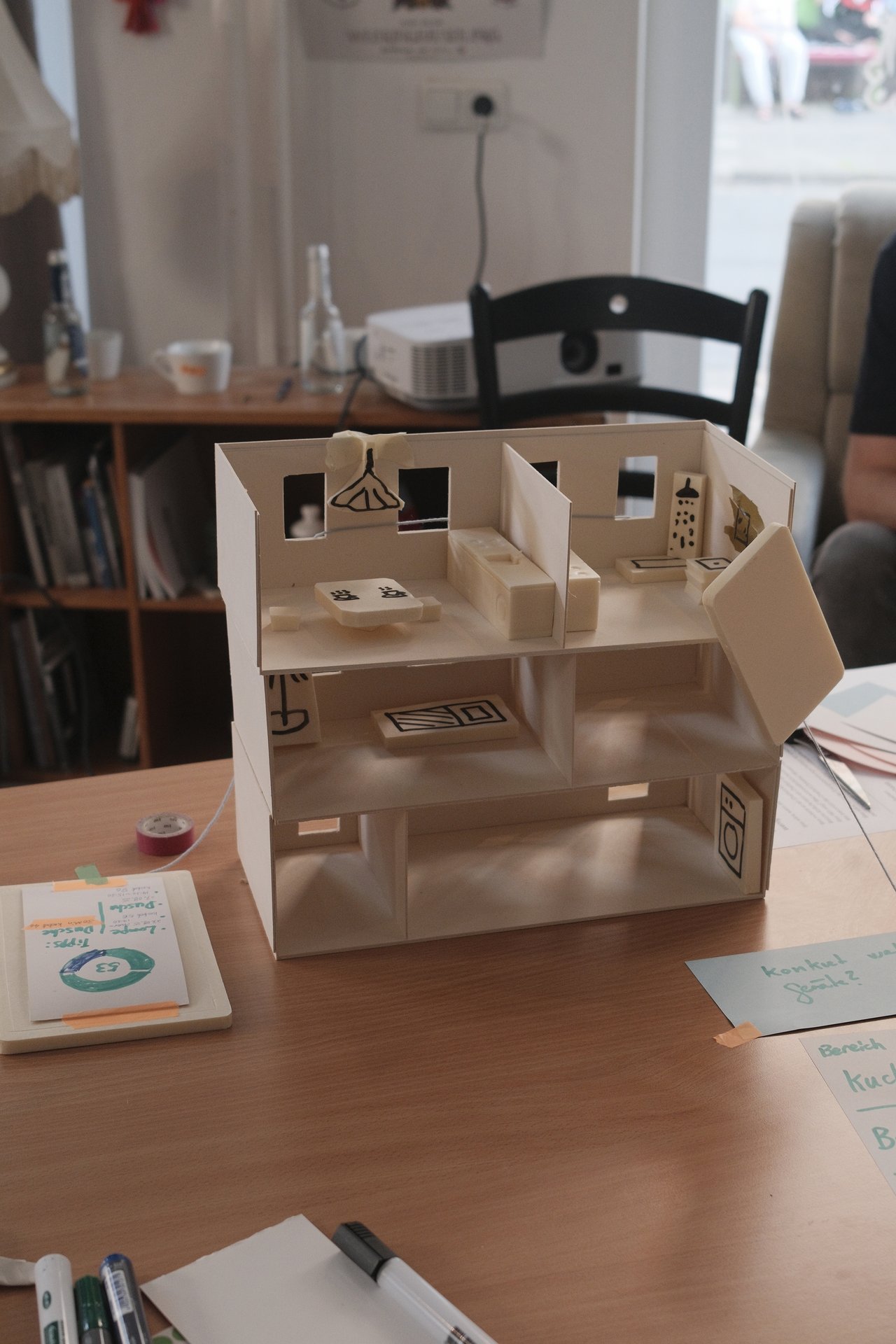 Ein kleines, gebasteltes Modellhaus befindet sich auf einem Tisch. Durch eine fehlende Hauswand zeigt sich die Raumaufteilung und Einrichtung des Hauses.