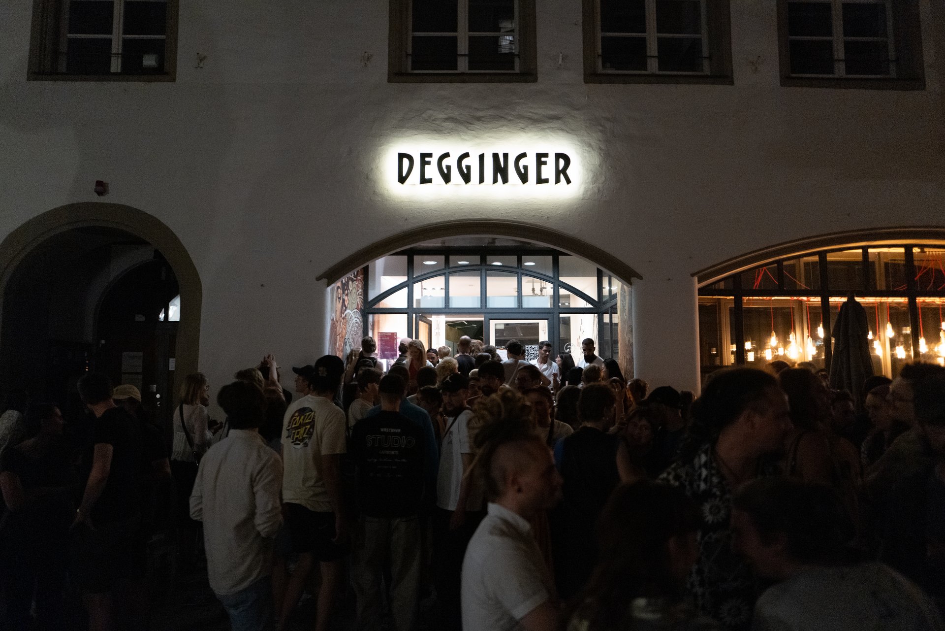 Viele junge Menschen befinden sich abends vor dem Eingang eines Gebäudes mit der Aufschrift "Degginger". 