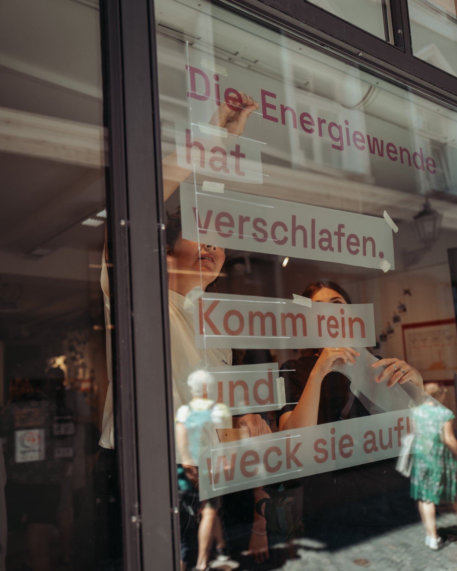 Ein Schaufenster, in welchem von innen zwei Frauen den Text "Die Energiewende hat verschlafen. Komm rein und weck sie auf." aufhängen.