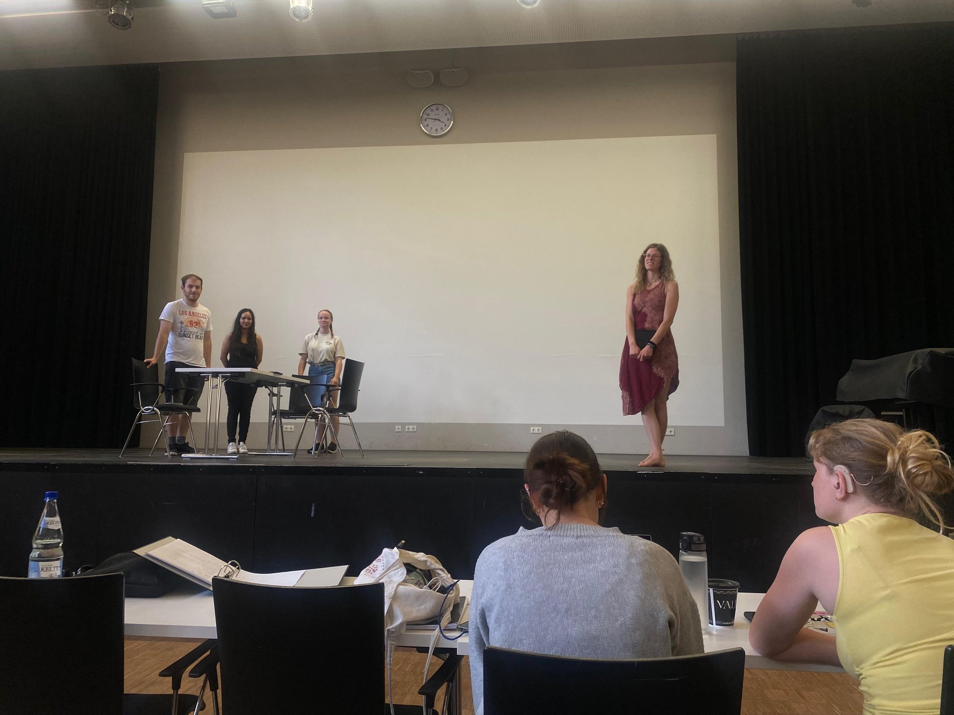 Vier Studierende stehen bei der Probe des Stückes auf der Bühne. Zwei weitere Personen sitzen an Tischen vor der Bühne.