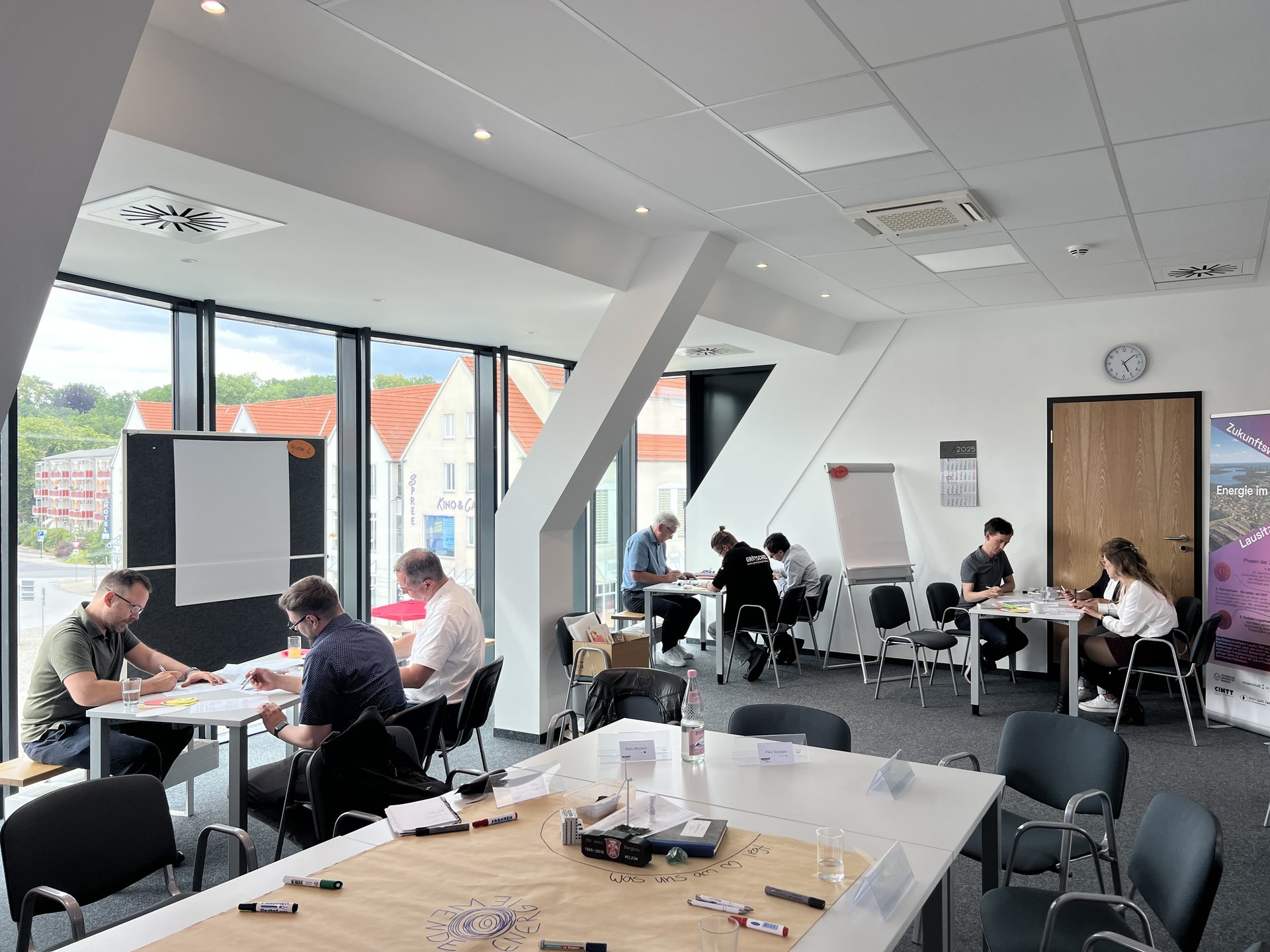 Es sitzen mehrere Kleingruppen an einzelnen Tischen und arbeiten konzentriert. Sie befinden sich in einem großen Büro.