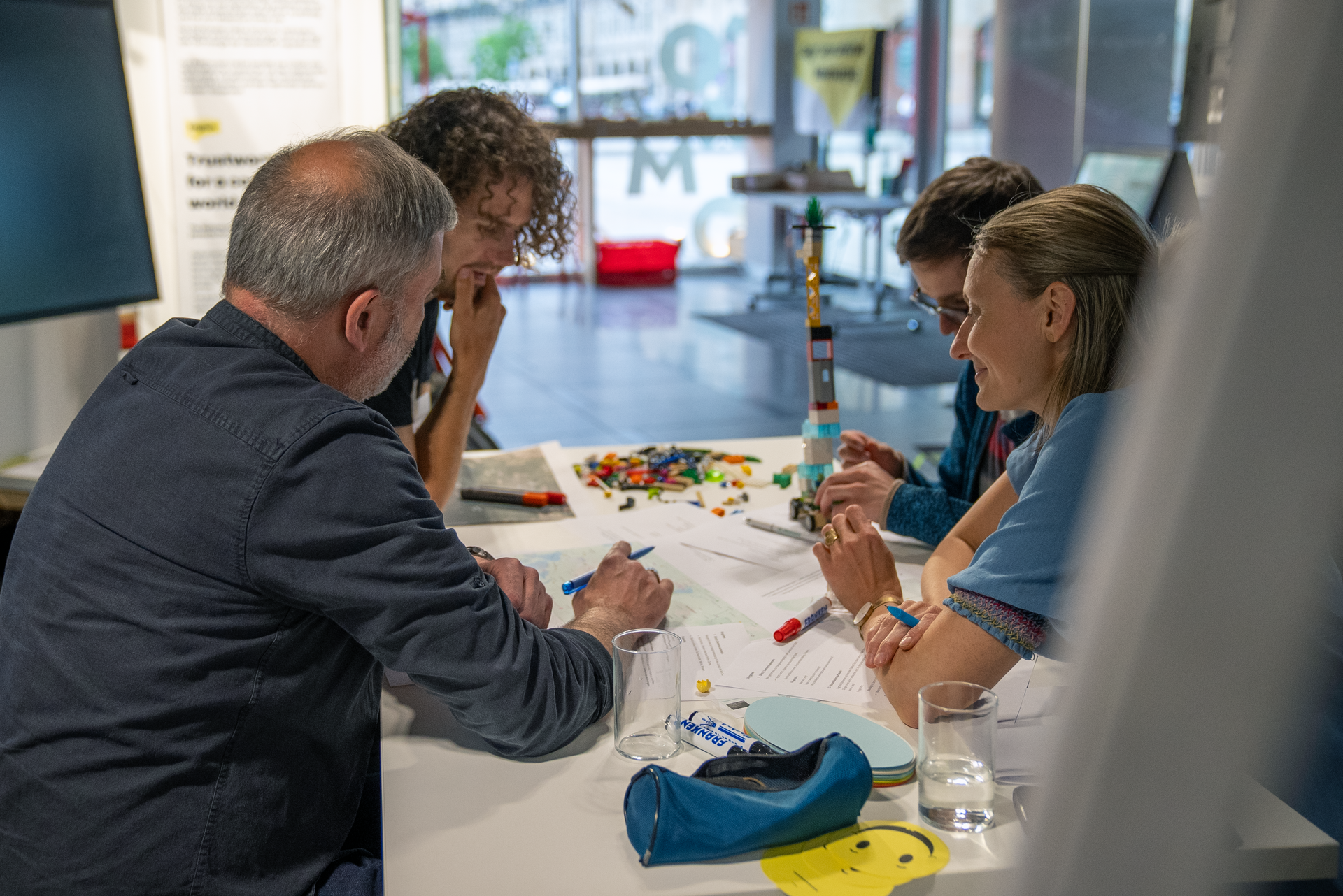 Vier Personen sitzen an einem Tisch. Sie sind über Arbeitsblätter und Stifte gebeugt. Eine Person hält sich nachdenklich an das Kinn.