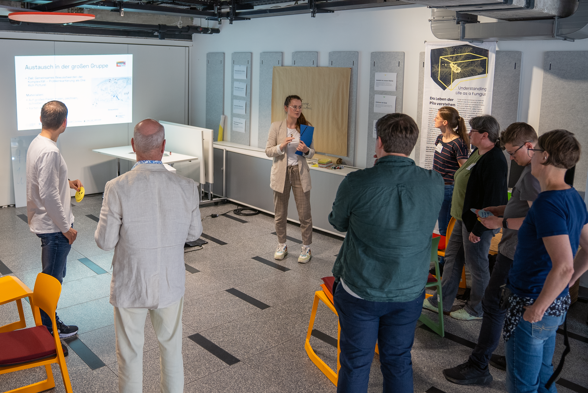 Eine Person hält einen Power-Point-Vortrag. Um sie herum stehen mehrere Personen in einem losen Halbkreis.