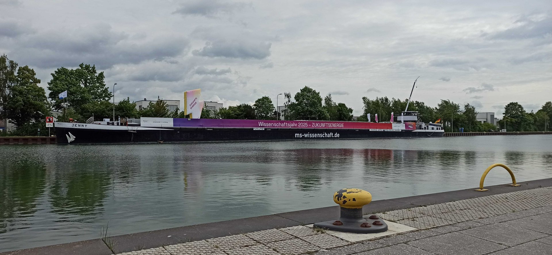 Schiff mit Aufschrift "Wissenschaftsjahr 2025 – ZUKUNFTSENERGIE" liegt am gegenüberliegenden Ufer an Ankerplatz