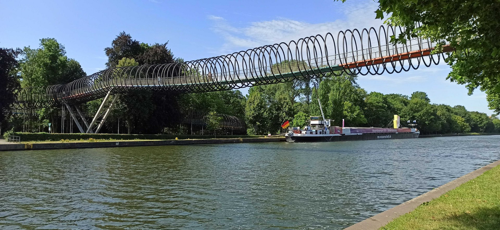 Fußgängerbrücke führt über einen Fluss, dahinter liegt ein Schiff mit Deutschlandfahne am gegenüberliegenden Ufer an einem Ankerplatz