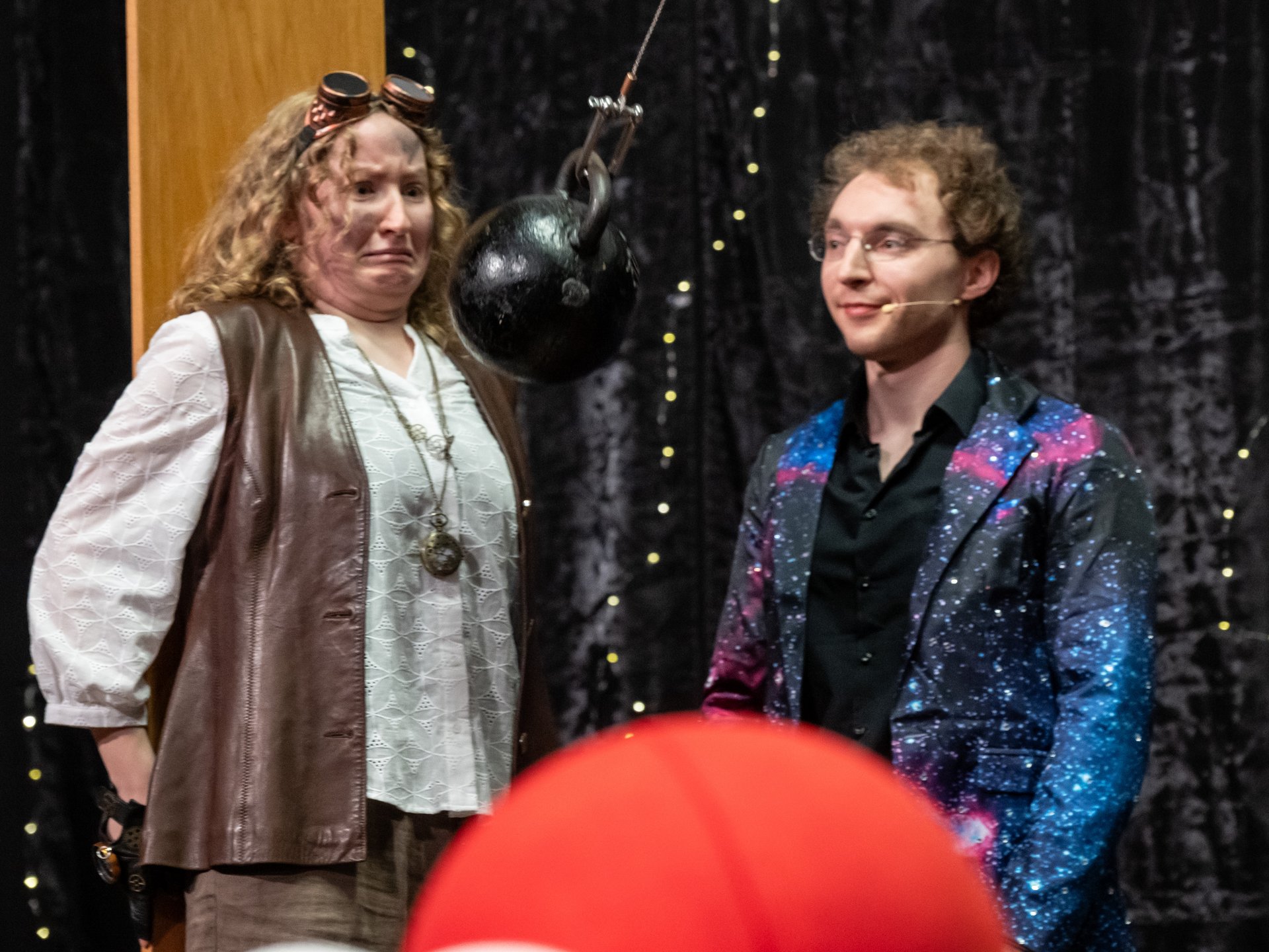 Verkleidete Schauspielerin links schaut erschreckt auf einen Kettleball, der in Richtung ihres Gesichts schwingt. Verkleideter Schauspieler rechts beobachtet das Geschehen.