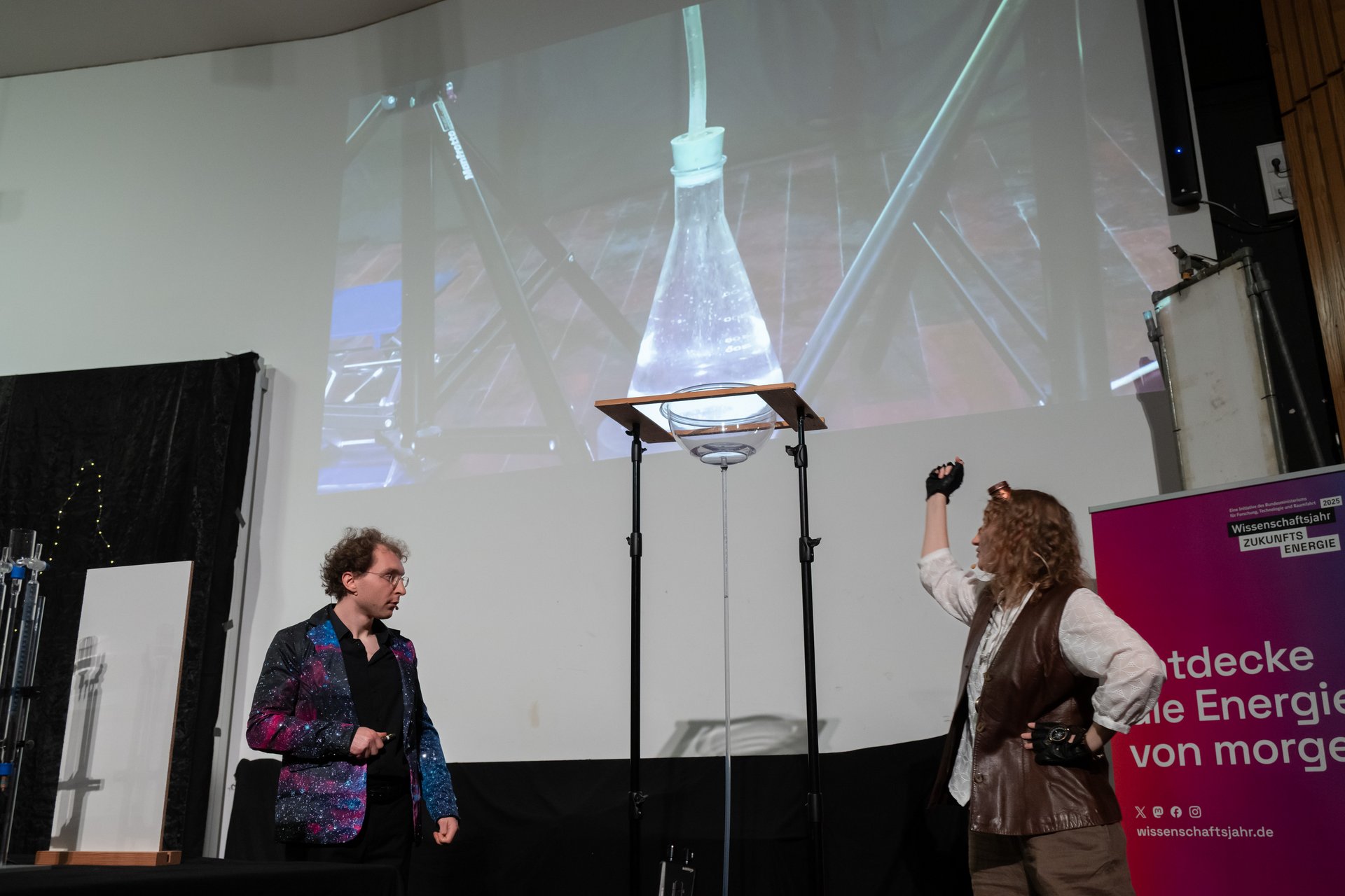 Verkleidete*r Schauspielerin und Schauspieler führen ein Live-Experiment auf der Bühne durch. An der Wand ist ein Bild eines Reagenzglases zu erkennen, das per Beamer übertragen wird.