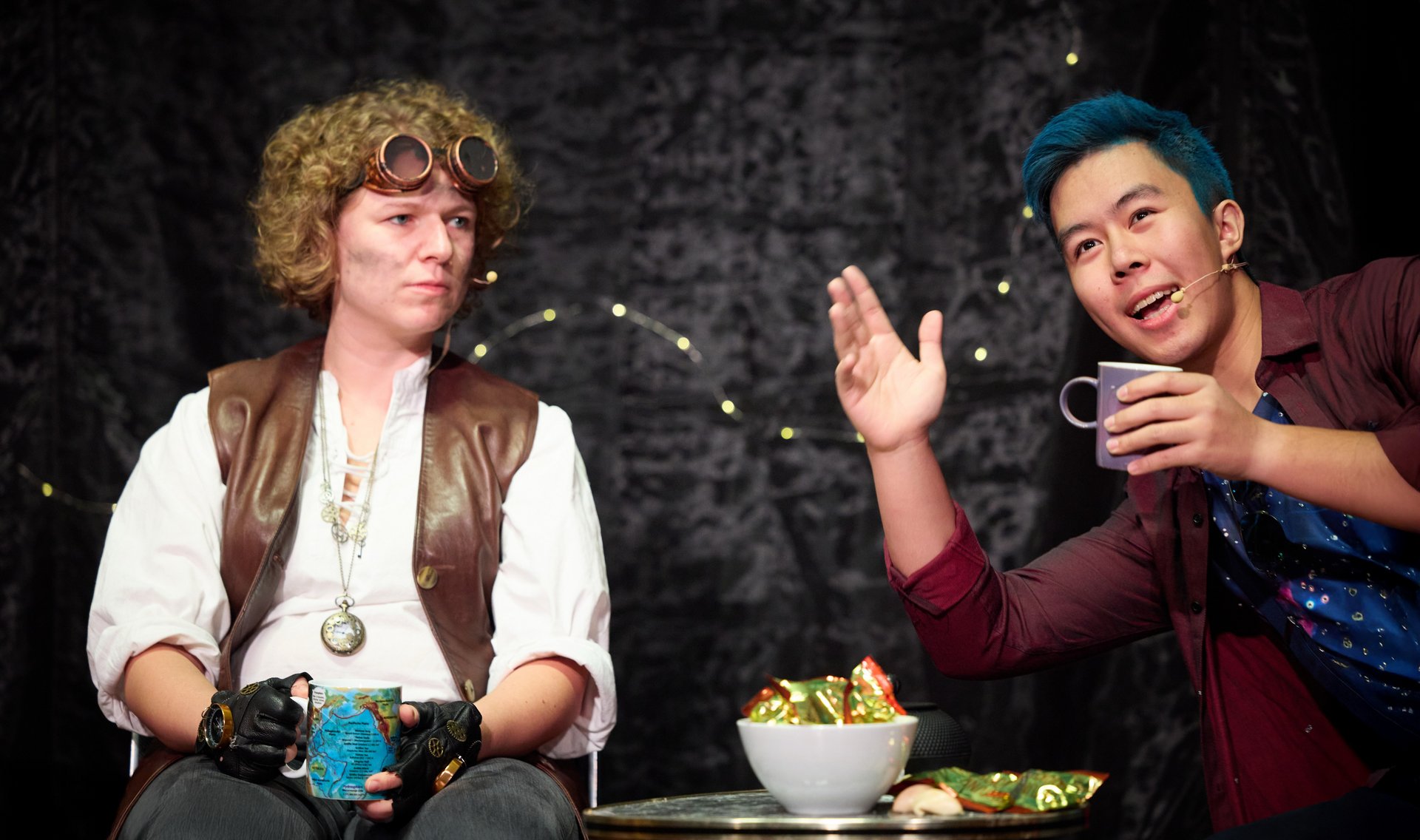 Verkleidete*r Schauspielerin und Schauspieler sitzen gemeinsam an einem kleine Tisch. Auf dem Tisch liegen Glückskekse. Die beiden halten jeweils eine Tasse in der Hand, während der Schauspieler dem Publikum etwas erzählt.