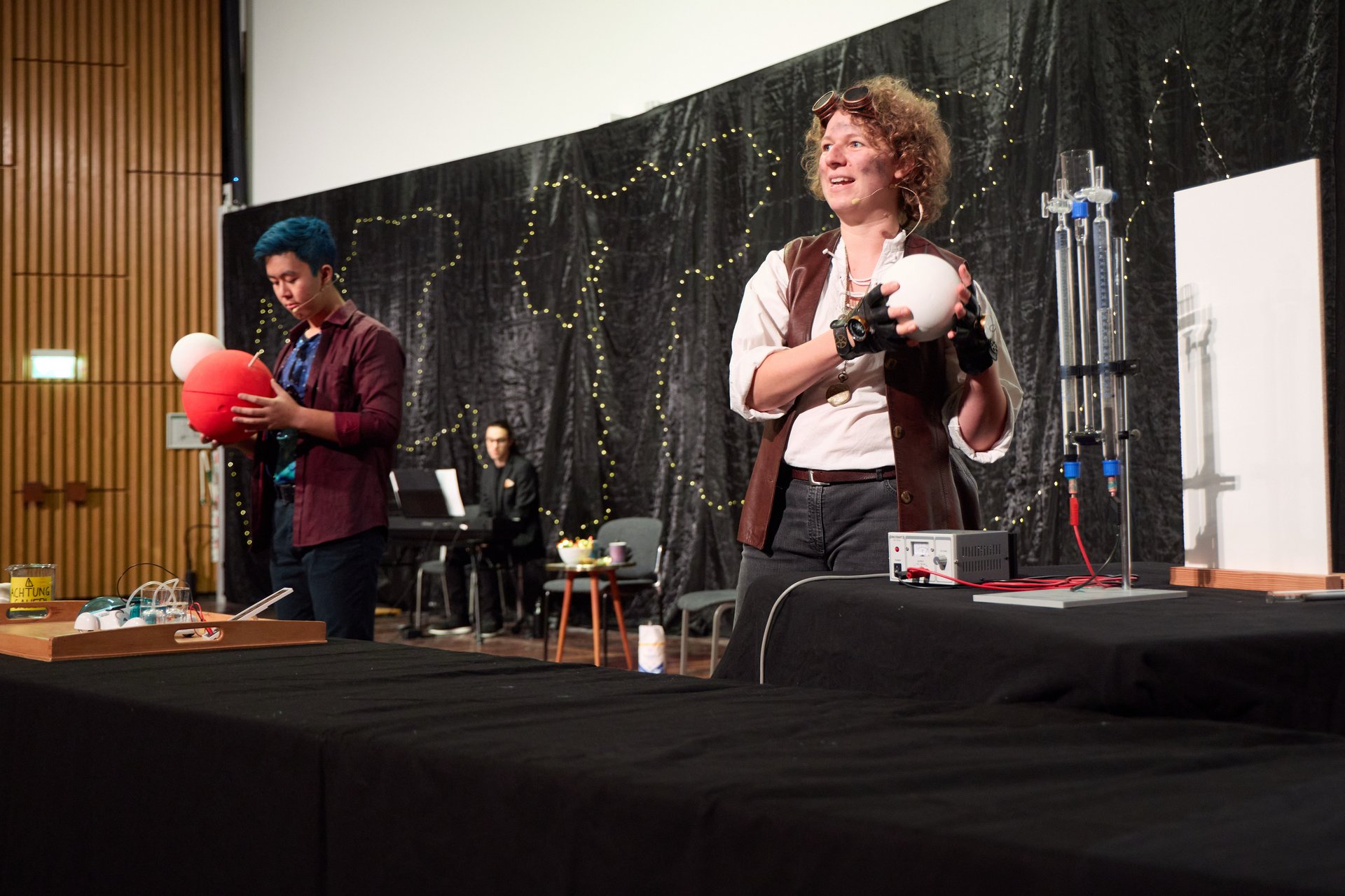 Verkleidete*r Schauspielerin und Schauspieler führen ein Live-Experiment durch und halten beide jeweils eine Kugel in der Hand. 