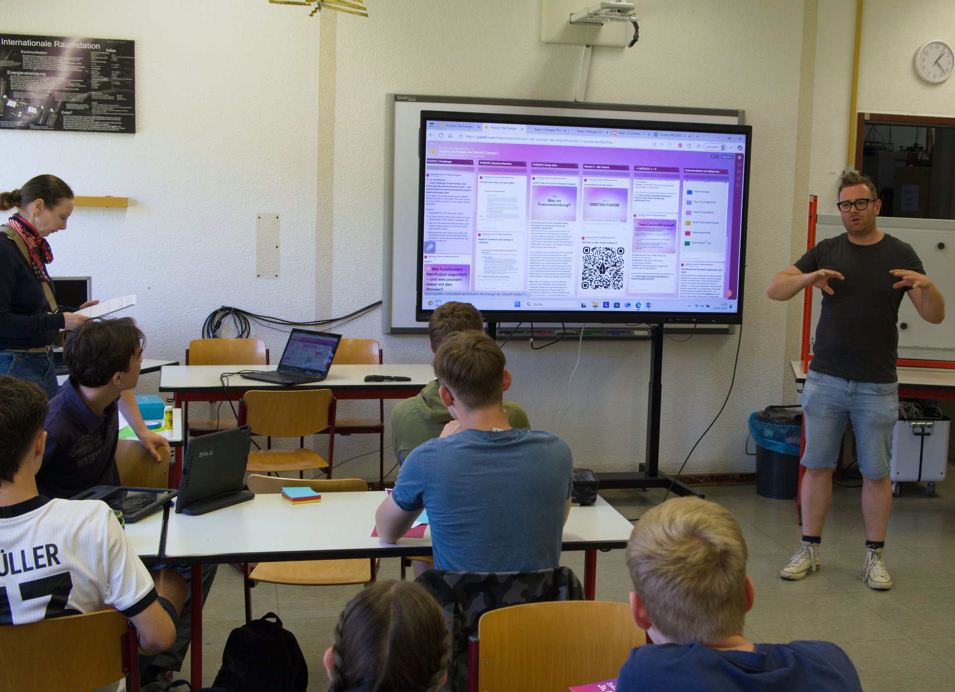 Person steht vor sitzenden Menschen in einem Klassenraum, im Hintergrund eine Präsentation.