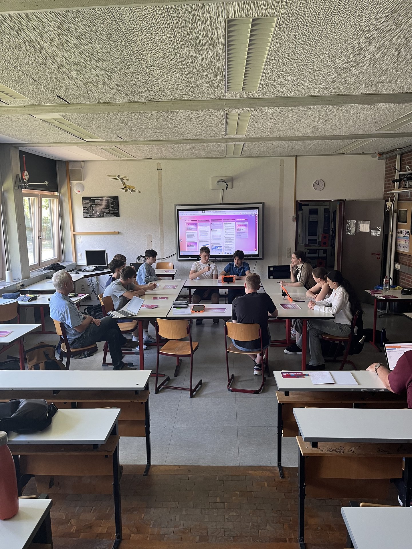 Elf Personen sitzen an zum Kreis zusammengestellten Tischen in einem Klassenraum.