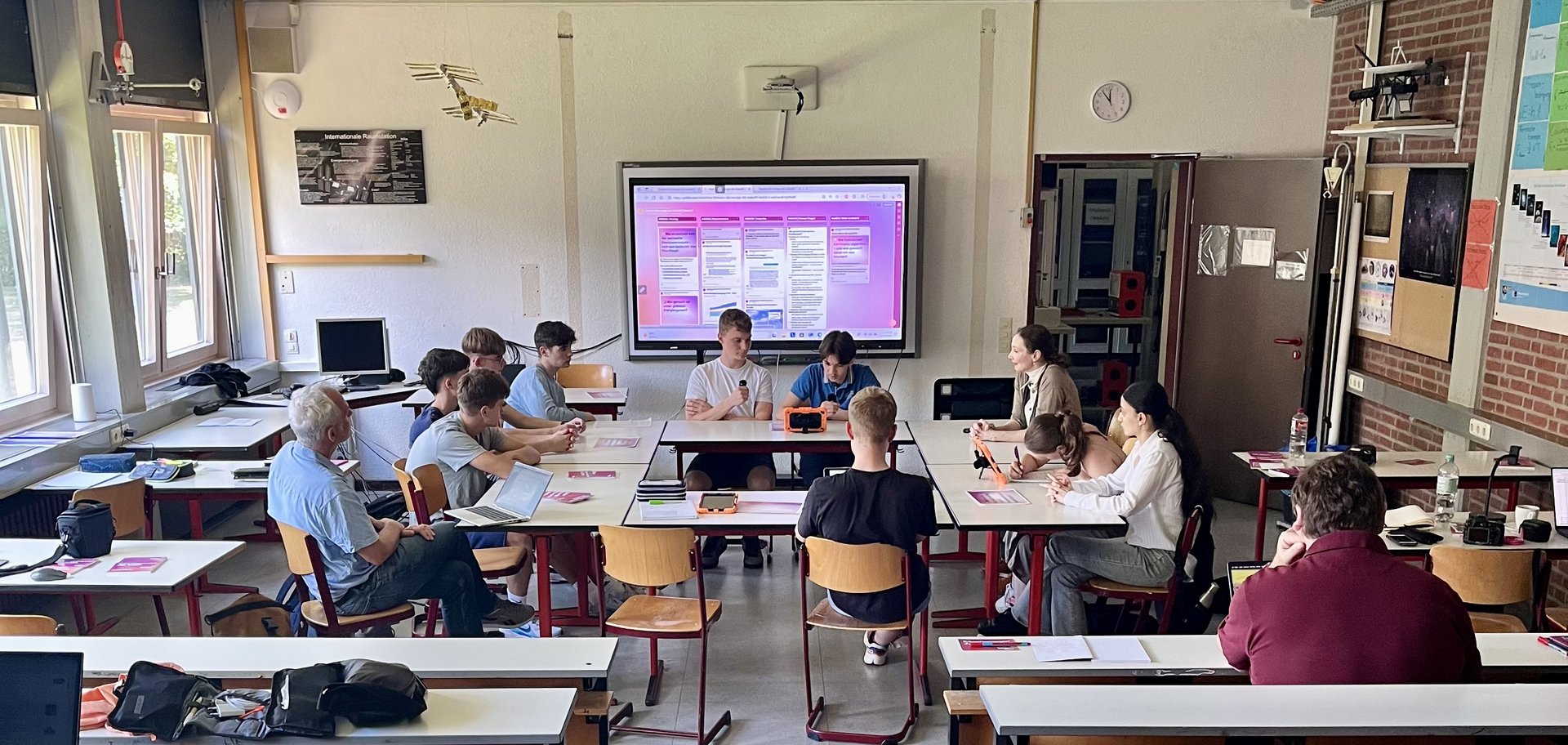 Elf Personen sitzen an zu Kreis zusammengestellten Tischen in einem Klassenraum.