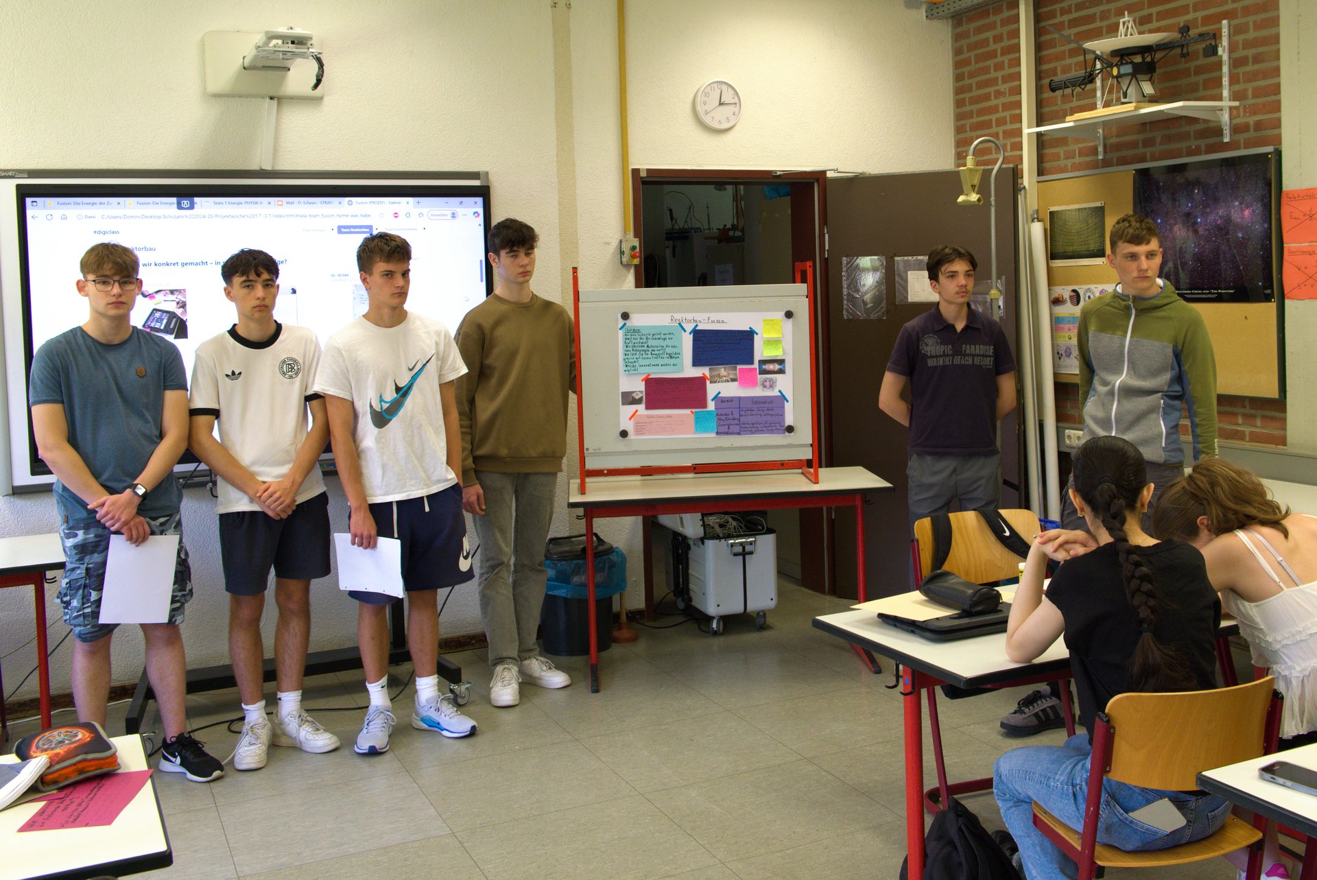 Sechs junge Personen stehen vorne im Klassenraum, an den Tischen im Raum sitzen weitere Personen.