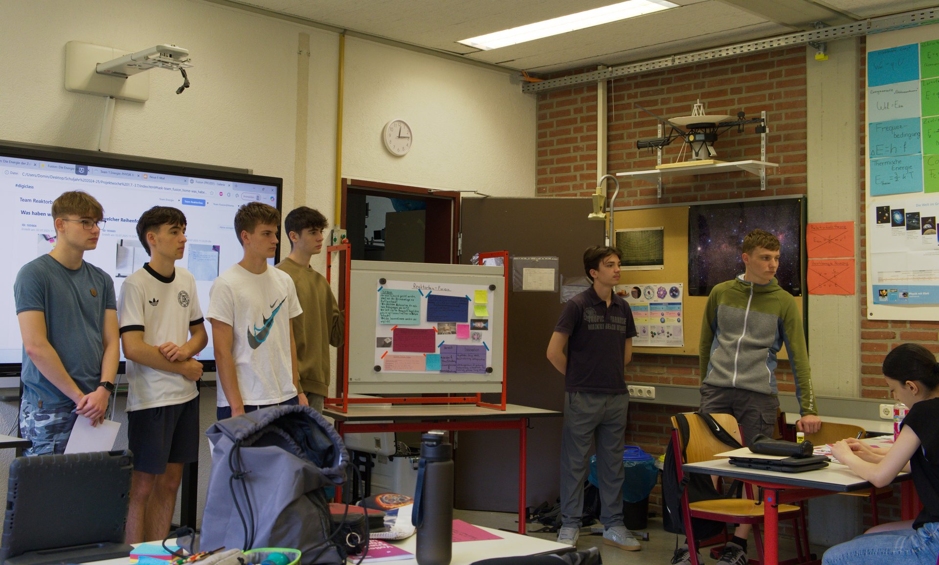 Sechs junge Personen stehen vorne im Klassenraum, an den Tischen im Raum sitzen weitere Personen.