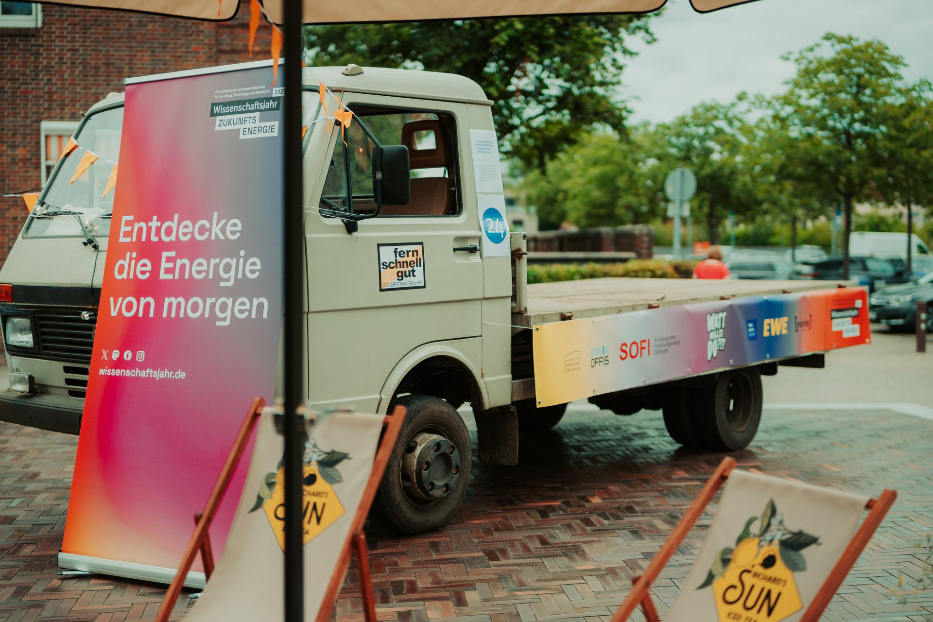 LKW mit leerer Ladefläche, davor ein Roll-up mit der Aufschrift "Entdecke die Energie von morgen".