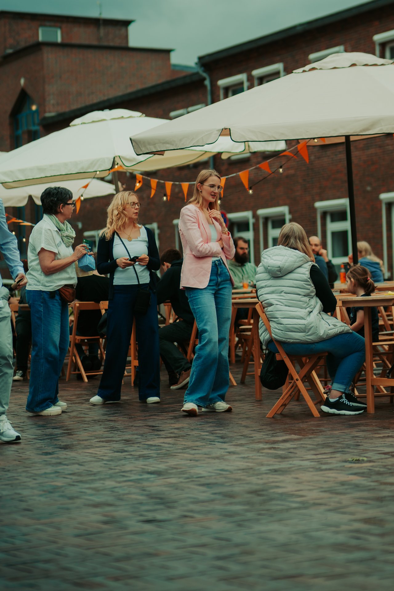 Mehrere Personen sitzen an Tischen unter Sonnenschirmen, drei Personen stehen vor den Tischen.
