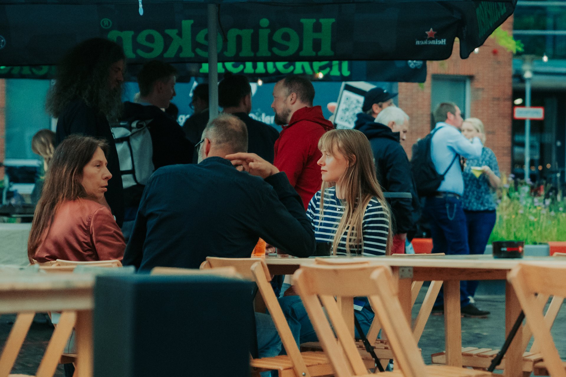 Drei Personen sitzen an Tischen auf einem Straßenfest und unterhalten sich, im Hintergrund stehen weitere Personen.