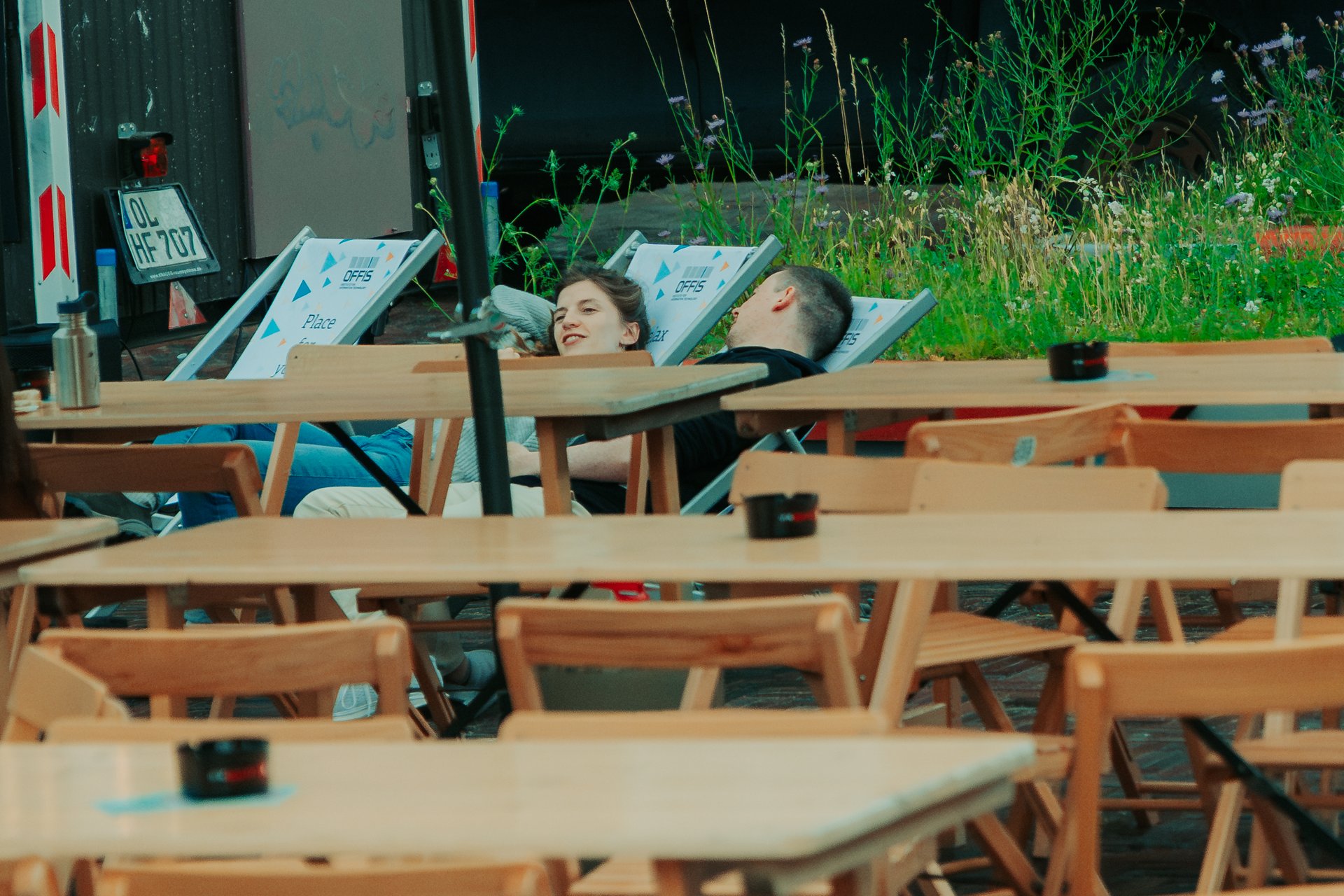 Mehrere Tische und Stühle, im Hintergrund eine Frau und ein Mann auf Liegestühlen.