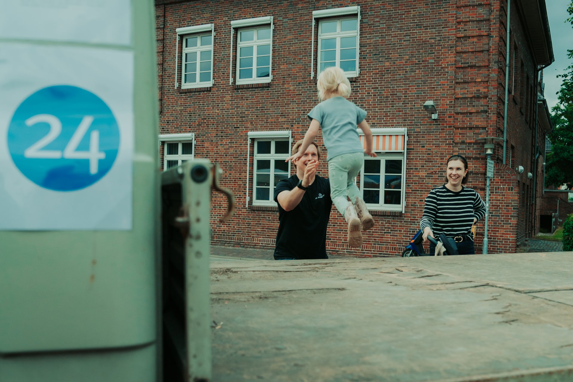 Kind springt von der Ladefläche eines Autos in die Arme eines Mannes, daneben steht eine Frau und grinst.
