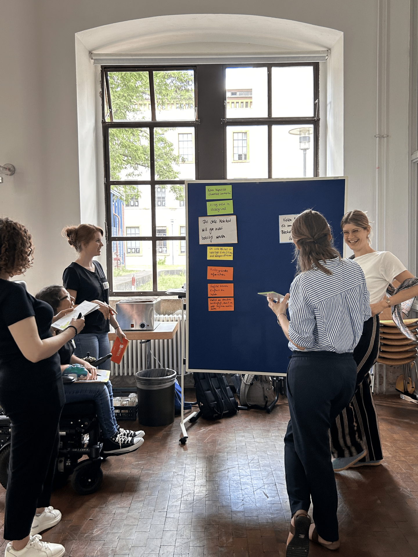 Eine Gruppe steht vor einer Pinnwand mit Zetteln und diskutiert.