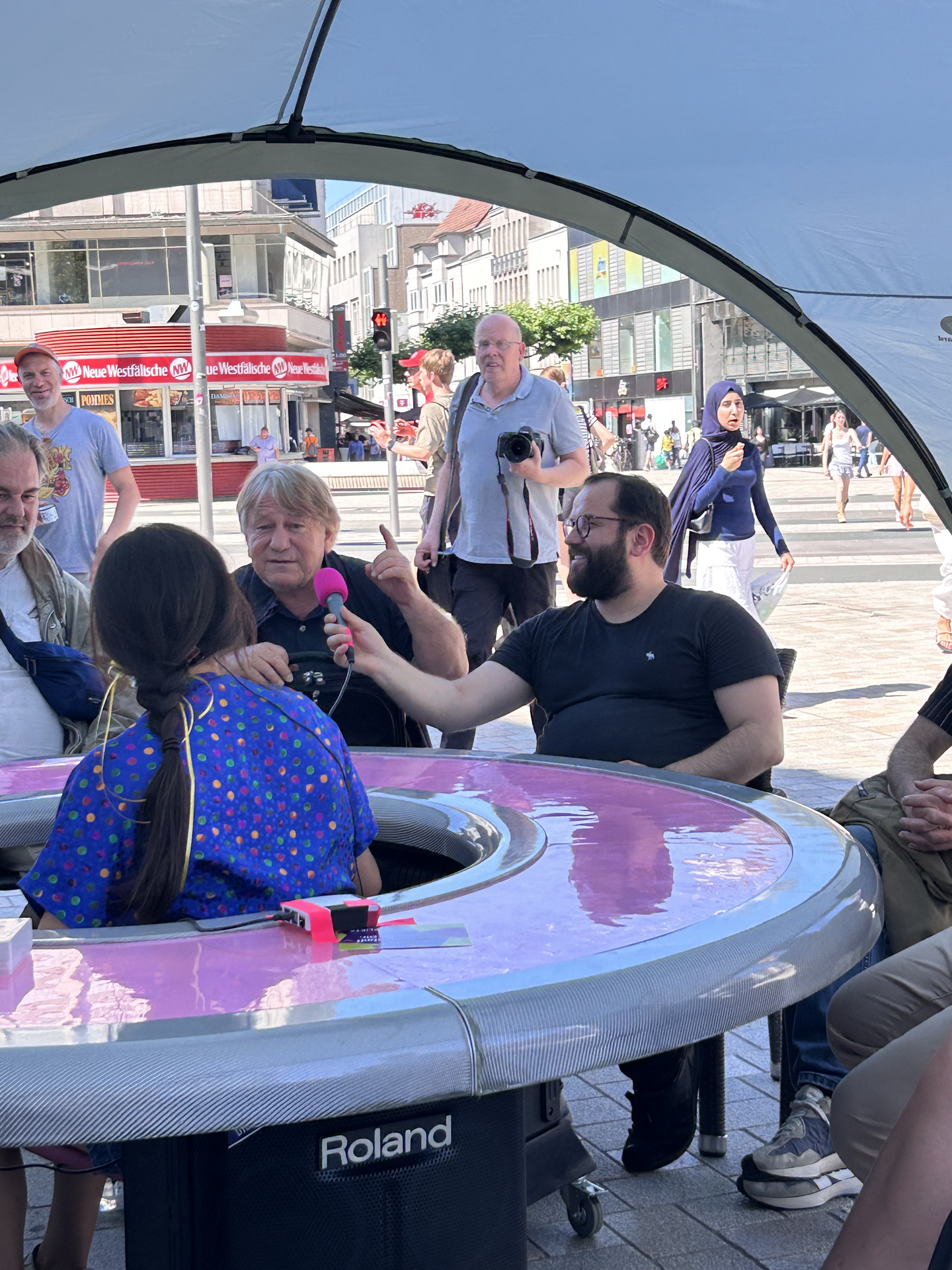 Ein Mann sitzt mit anderen am runden Tisch und spricht in ein Mikrofon.