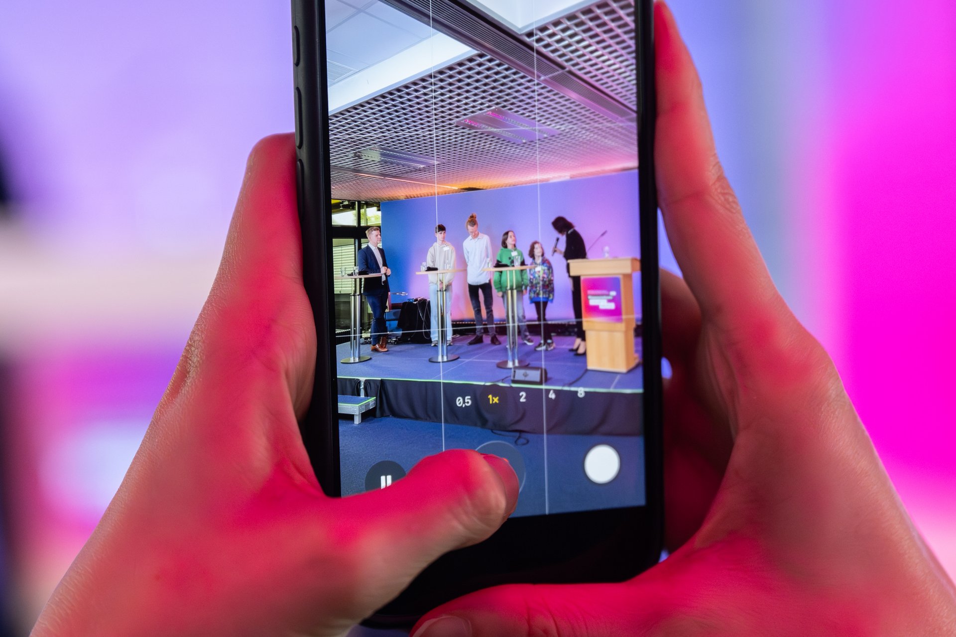 Zwei Hände halten ein Handy, auf dem mehrere Menschen auf einer Bühne abgebildet sind.