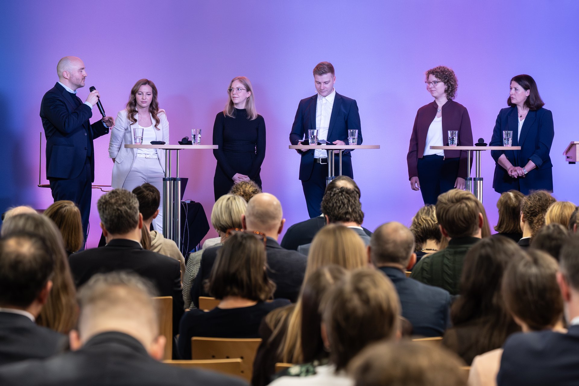 Sieben Menschen stehen im Hintergrund auf einer Bühne, im Vordergrund sitzt das Publikum.