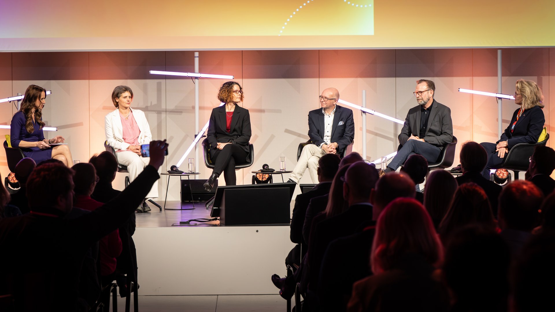Sechs Personen sitzen im Halbkreis auf Stühlen auf einer Bühne.