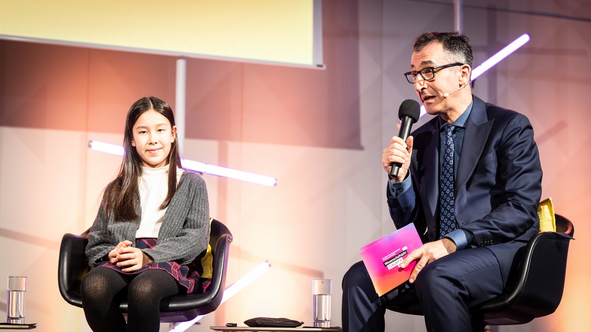 Bundesminister für Bildung und Forschung Cem Özdemir sitzt neben einem Mädchen und spricht in ein Mikrofon.
