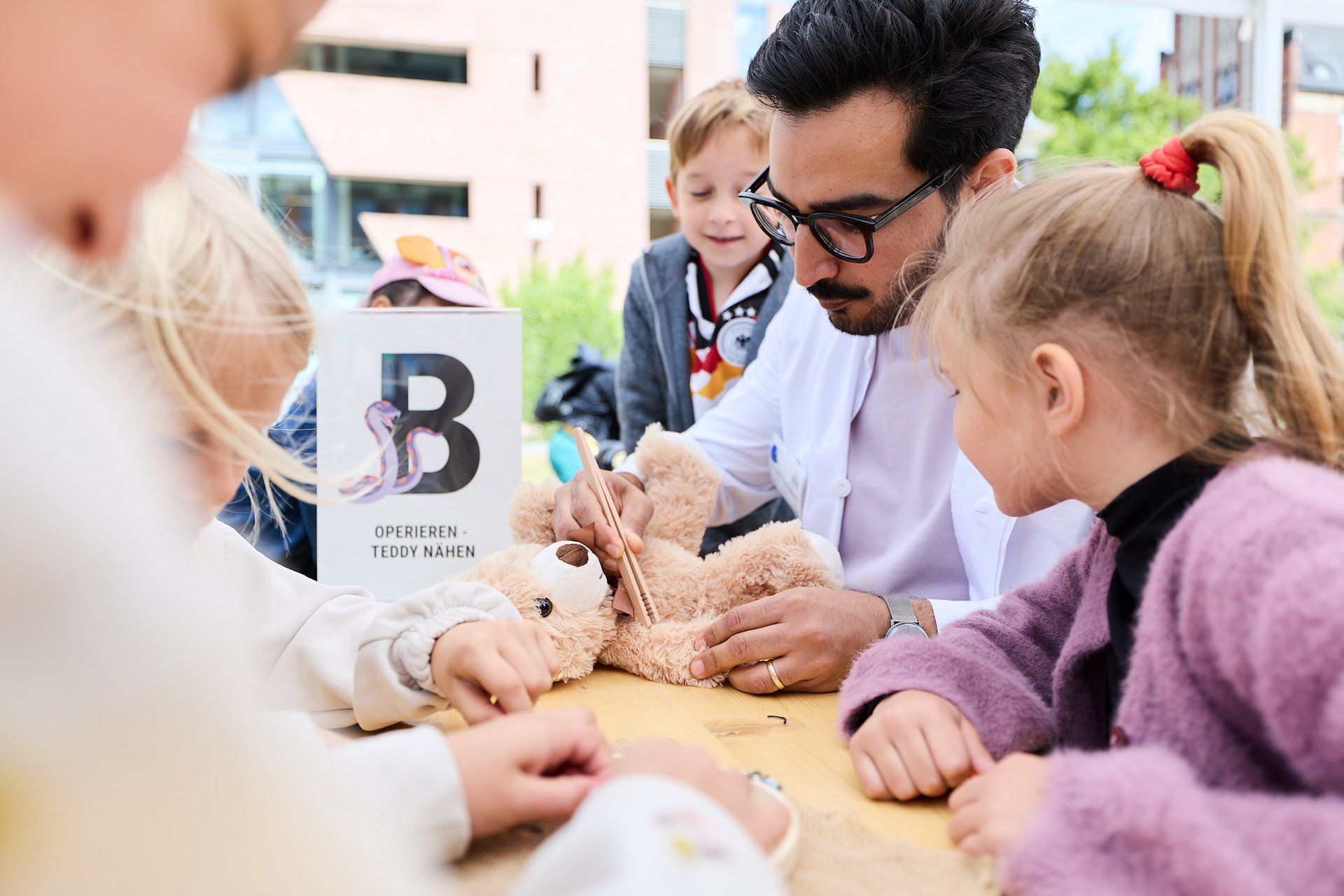 Dr. med. Nibras Naami berührt einen auf einem Tisch liegenden Teddybär mit einer Zange, fünf Kinder stehen um ihn herum.