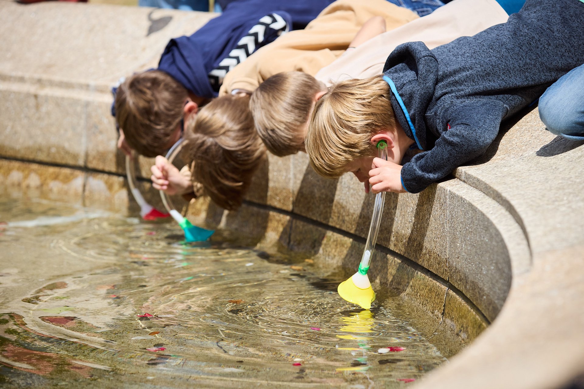 Vier Kinder beugen sich über den Rand eines Brunnens und halten Trichter ins Wasser, an denen Schläuche befestigt sind, die sie sich ans Ohr halten.
