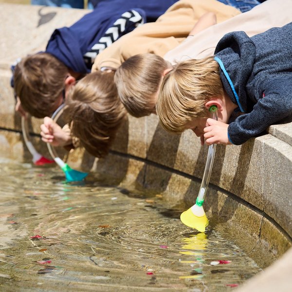 Vier Kinder beugen sich über den Rand eines Brunnens und halten Trichter ins Wasser, an denen Schläuche befestigt sind, die sie sich ans Ohr halten.
