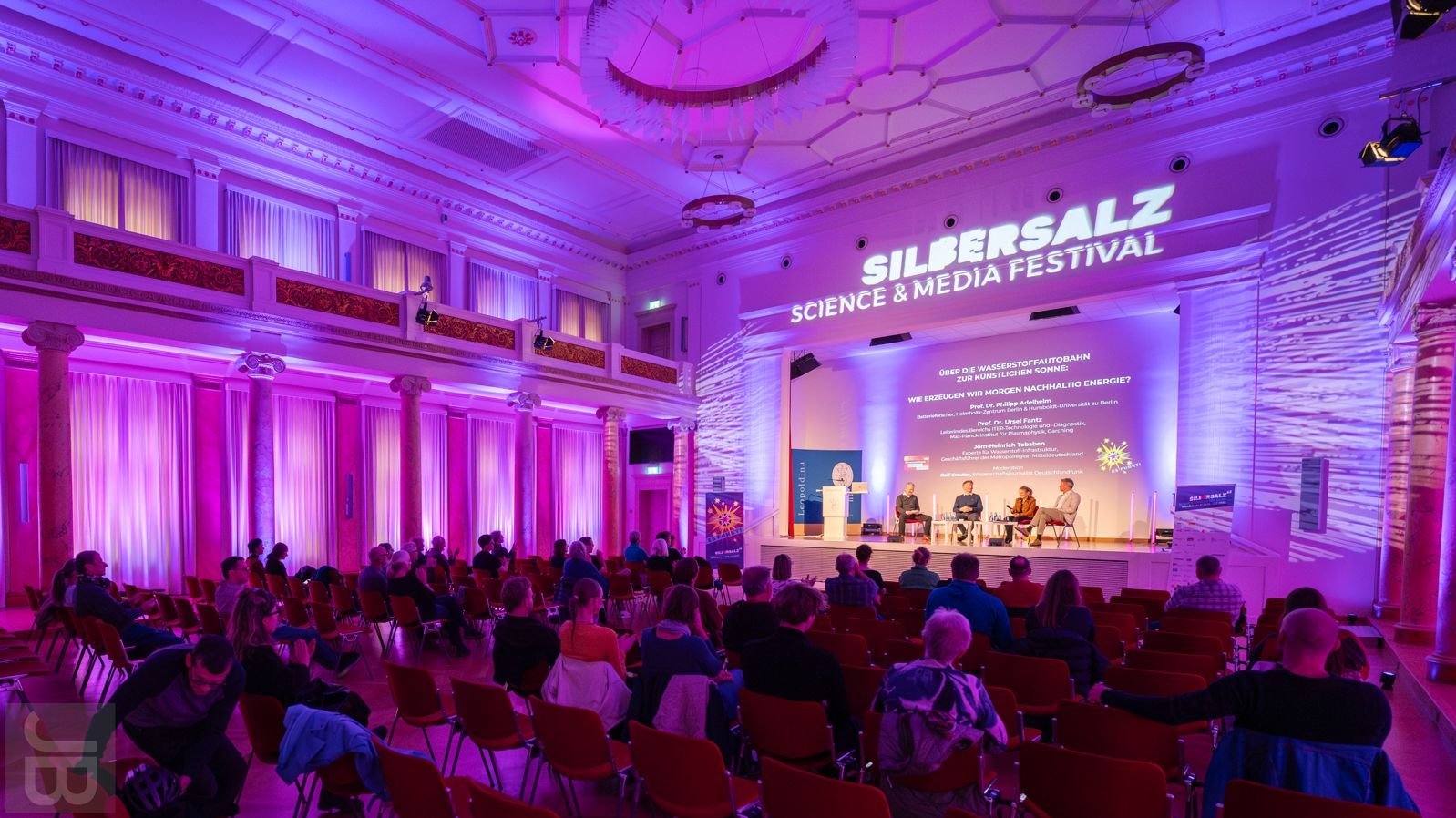 Vier Personen sitzen auf Stühlen auf einer beleuchteten Bühne vor einem Publikum