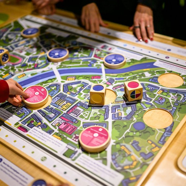 Ein Holzspielbrett, das eine Stadt zeigt. Darauf liegen bunte Würfel und Scheiben.
