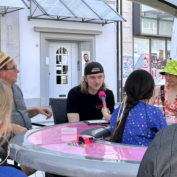 Eine junge Person sitzt mit anderen am runden Tisch und spricht in ein Mikrofon.