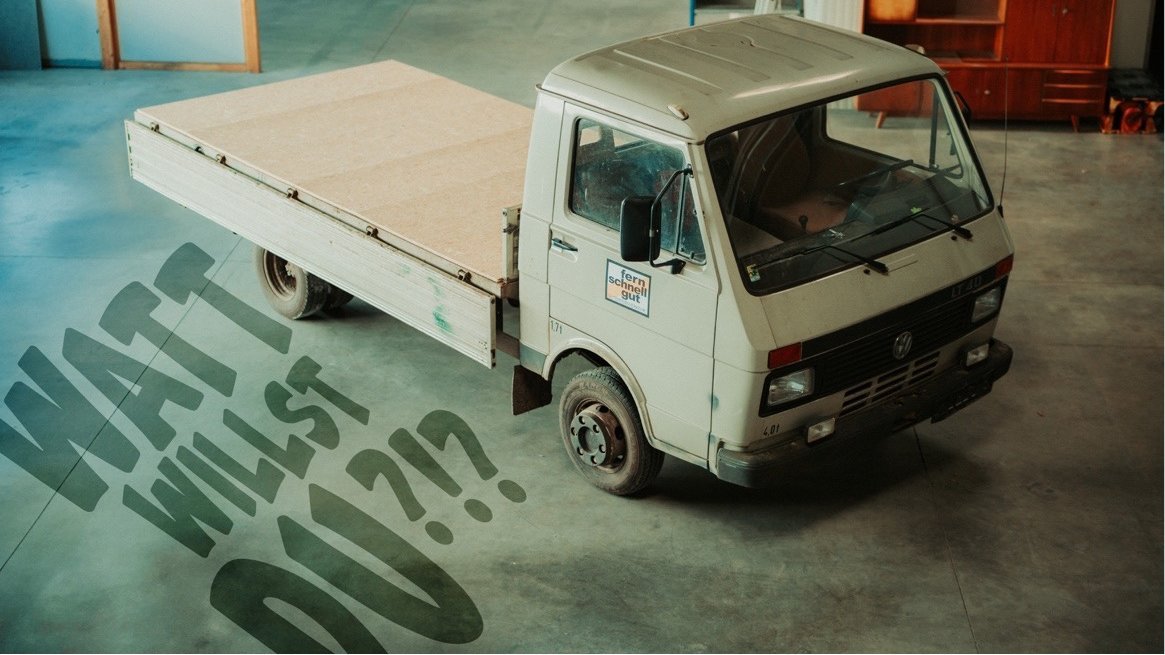 Ein alter, cremefarbener Pritschenwagen steht in einer Halle mit Betonboden. Auf dem Boden links vom Fahrzeug steht in großen, aufgemalten Buchstaben: „WATT WILLST DU?!?“. In der Halle befinden sich außerdem Gegenstände einer Werkstatt.