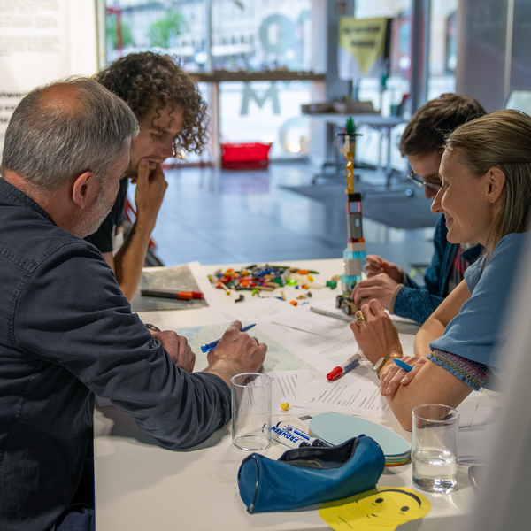 Vier Personen sitzen an einem Tisch. Sie sind über Arbeitsblätter und Stifte gebeugt. Eine Person hält sich nachdenklich an das Kinn.