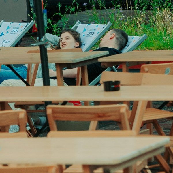 Mehrere Tische und Stühle, im Hintergrund eine Frau und ein Mann auf Liegestühlen.