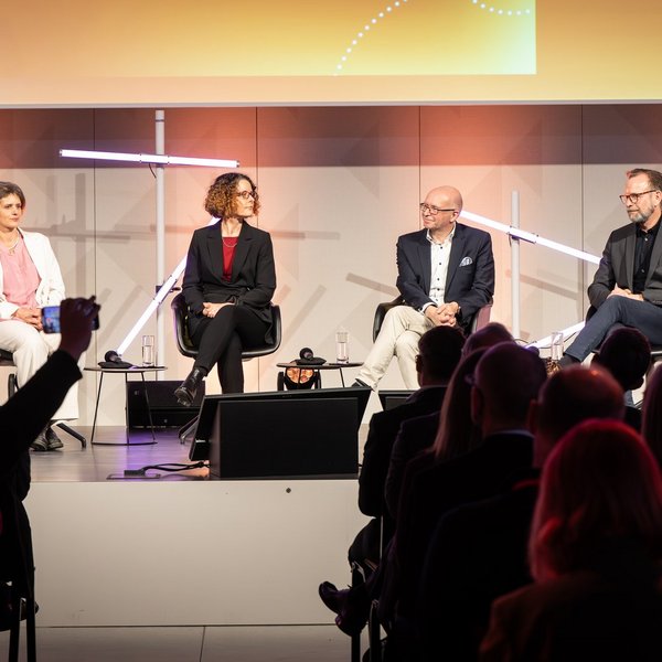 Sechs Personen sitzen im Halbkreis auf Stühlen auf einer Bühne.